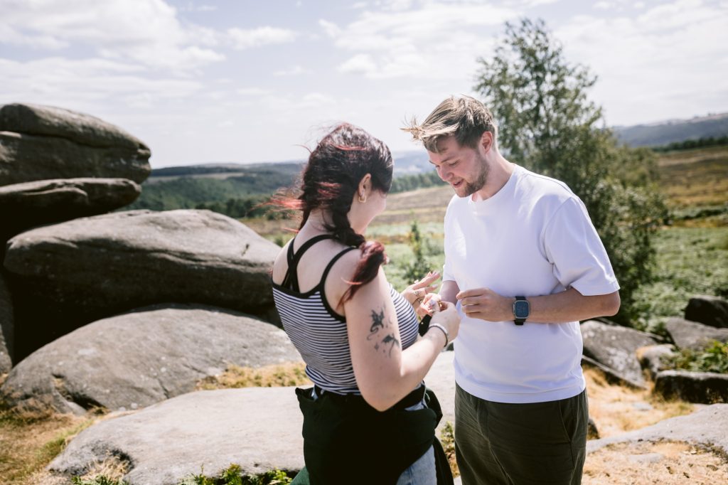 Two people stand outdoors by large rocks at Owler Tor in the Peak District; one is placing a ring on the other's finger during a surprise proposal on a sunny day.