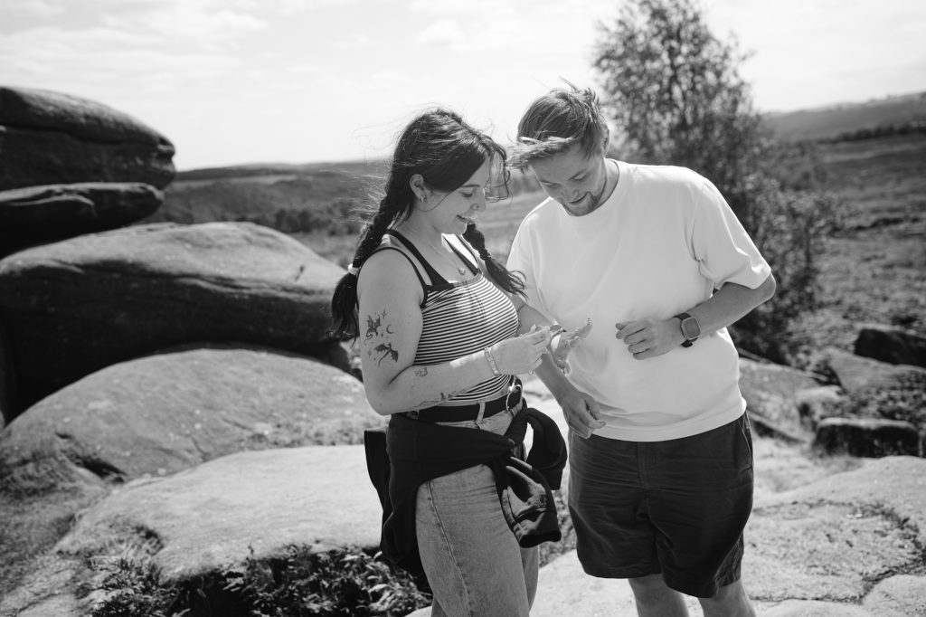Two people stand outdoors on the rocky terrain of Owler Tor in the Peak District, looking down at an object in one person's hands. They appear relaxed and are dressed casually for warm weather, perhaps caught up in the excitement of a surprise engagement.
