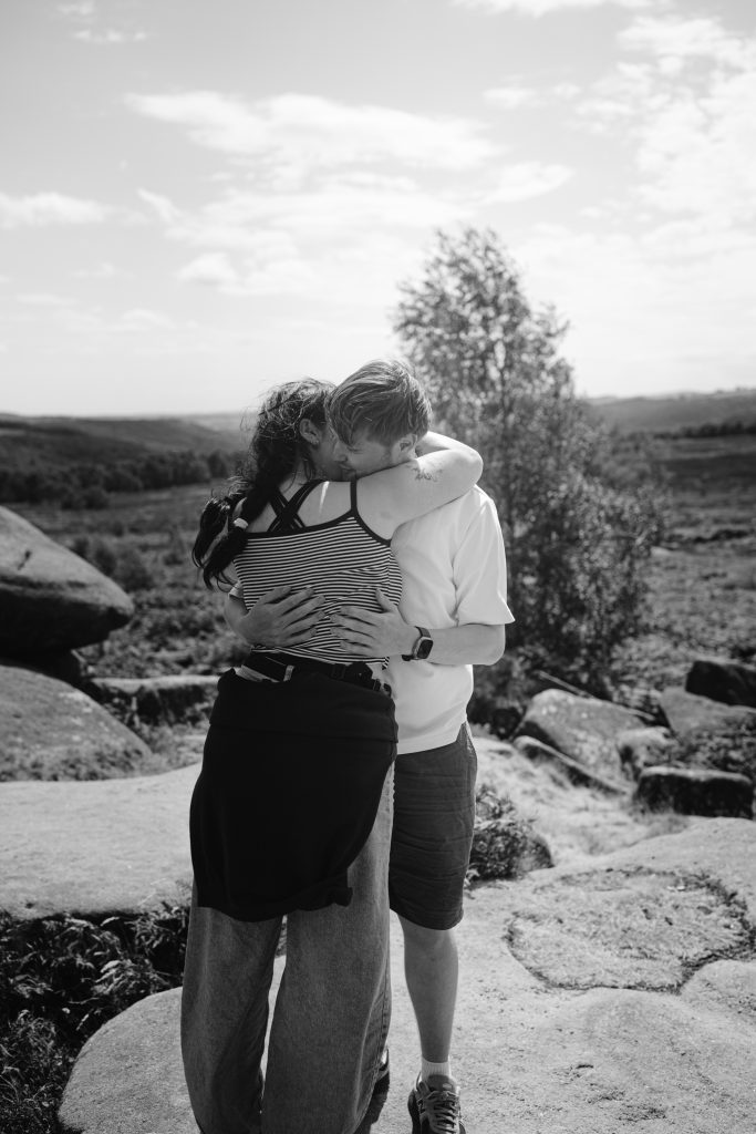 Two people embrace and kiss outdoors on a rocky landscape at Owler Tor, beneath open sky and surrounded by trees in the Peak District, captured in black and white—a moment that hints at a surprise proposal amid nature’s beauty.
