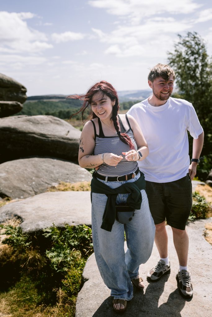 Two people stand on large rocks outdoors at Owler Tor in the Peak District, smiling on a windy day. The woman holds something in her hands—a hint of a surprise proposal. Trees and distant hills are visible under a partly cloudy sky.