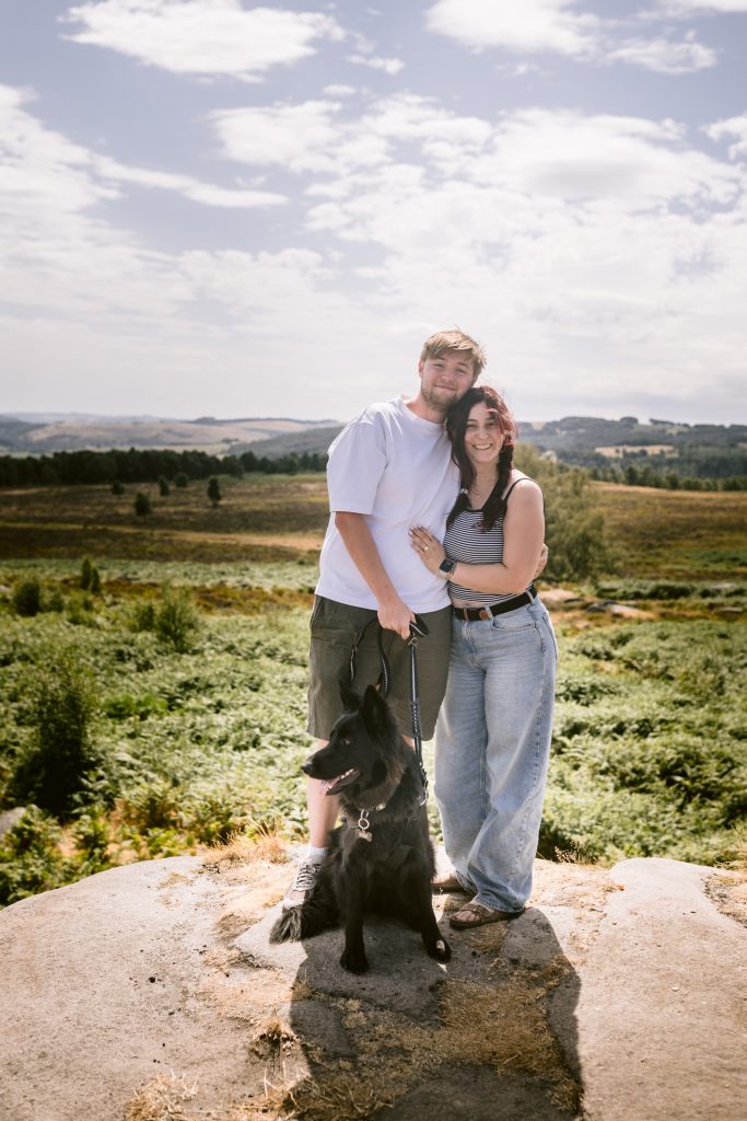 A couple stands on a rocky hilltop at Owler Tor in the Peak District with a black dog on a lead. Rolling green fields and trees stretch into the distance under a partly cloudy sky, setting the perfect scene for a surprise proposal.