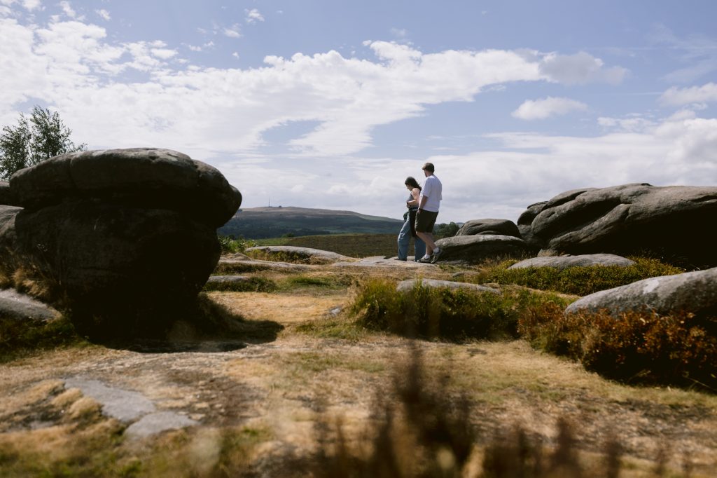 Two people walk among large rocks and grass under a partly cloudy sky, with hills visible in the background, exploring the striking landscape of Owler Tor in the Peak District.