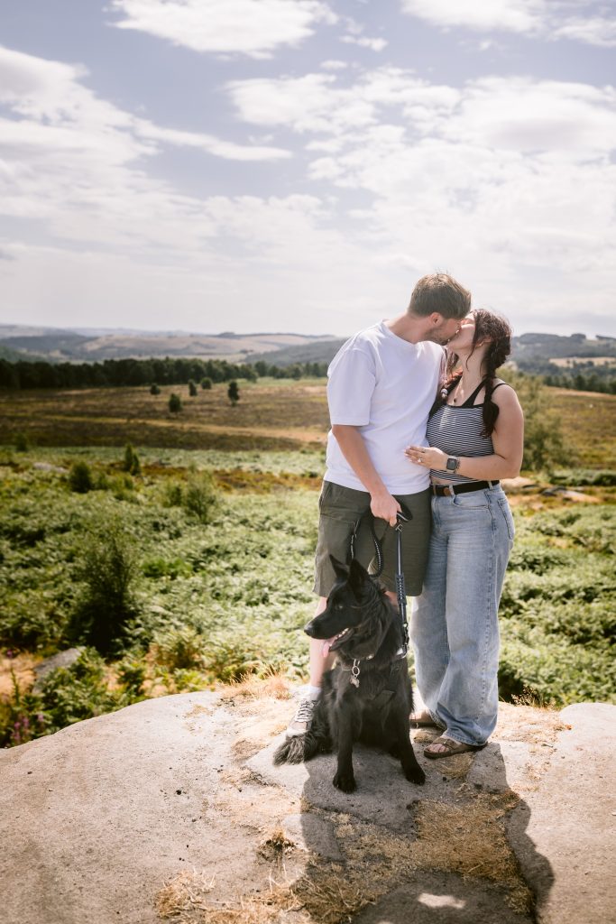 A man and woman stand on a rock in a grassy Peak District landscape near Owler Tor, the man kissing the woman's forehead in a tender moment, with a black dog sitting on a lead in front of them—capturing the magic of their surprise proposal.