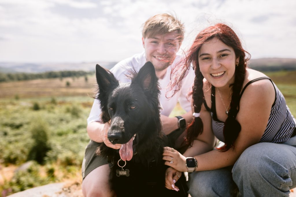 Two people smile at the camera outdoors, sitting close together with a large black dog. Set against the open field and sky of the Peak District near Owler Tor, this heart-warming moment could set the perfect scene for a surprise proposal.
