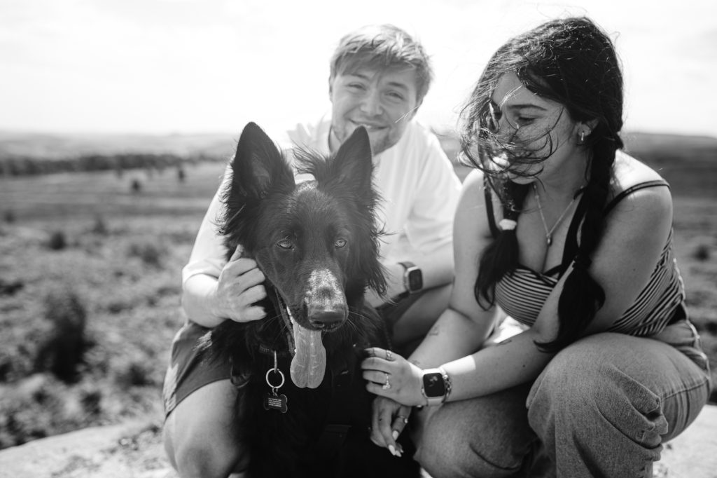 Two people crouch outdoors next to a large, long-haired dog. The dog faces the camera with its tongue out. The background shows open fields under a bright sky in the beautiful Peak District—a perfect setting for adventure and memories.