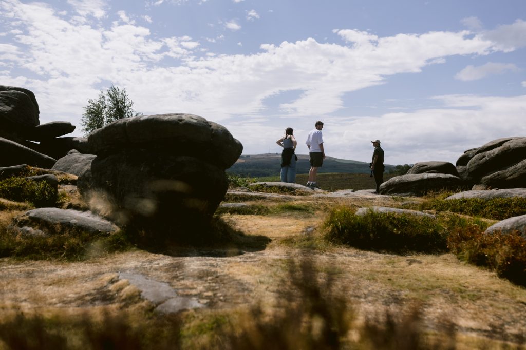 Three people stand on a rocky landscape beneath a partly cloudy sky at Owler Tor in the Peak District, with large boulders and distant hills visible in the background.