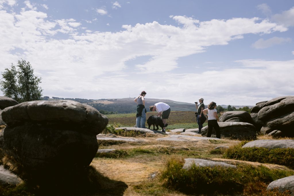Five people and a dog stand on the rocky landscape of Owler Tor in the Peak District under a partly cloudy sky, with distant hills visible in the background, capturing the excitement of a surprise engagement.