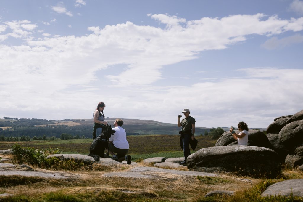 Four people and a large black dog stand and sit on rocky terrain at Owler Tor, with open fields and hills of the Peak District in the background under a partly cloudy sky.
