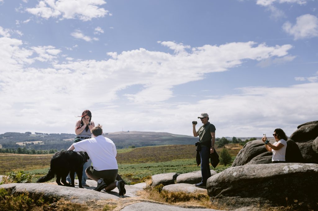 Four people and a black dog stand on rocks in the rural landscape of the Peak District, near Owler Tor; two people take photos whilst the others interact with the dog under a partly cloudy sky.