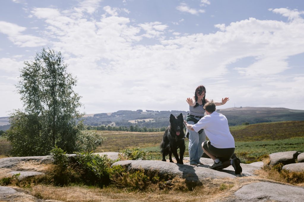 A person kneels and proposes to another person with a dog nearby on a rocky landscape at Owler Tor in the Peak District, under a partly cloudy sky—a perfect setting for a surprise engagement.
