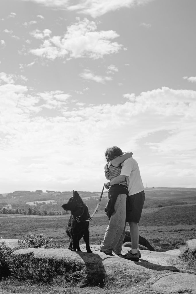 Two people embrace on a rocky hilltop at Owler Tor whilst a black dog on a lead stands nearby, looking away. The landscape features open fields and a cloudy sky, capturing the magic of a surprise proposal in the heart of the Peak District.