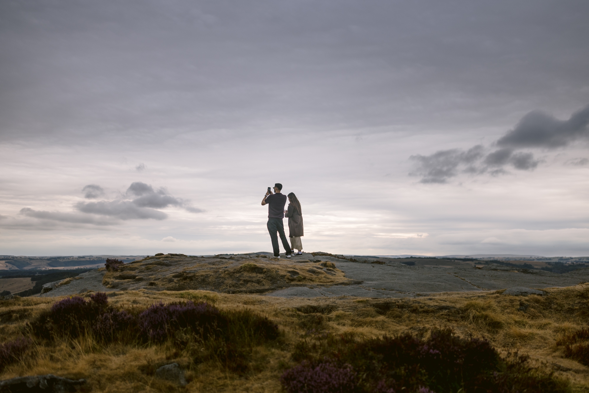 Two people stand together on a rocky hilltop under a cloudy sky at Curbar Edge, facing the horizon. One person appears to be using binoculars while the other stands close beside them—perhaps capturing the perfect moment for a Curbar Edge proposal.