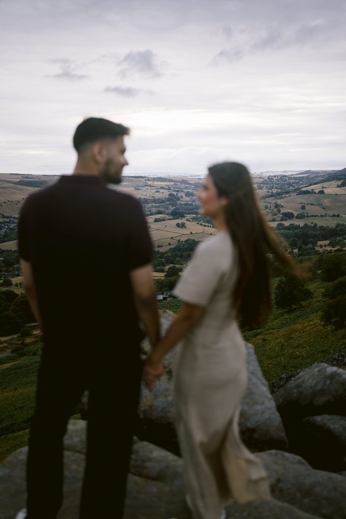 Two people stand on rocks holding hands, looking out over a hilly landscape with fields and a cloudy sky in the distance, capturing the magic of a Curbar Edge proposal.