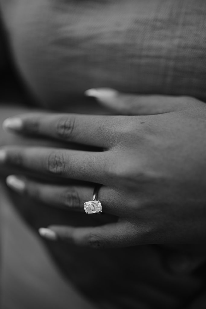 A close-up, black-and-white photo of a hand resting on fabric, wearing a square-cut diamond ring on the ring finger—a timeless image evocative of a Curbar Edge proposal.