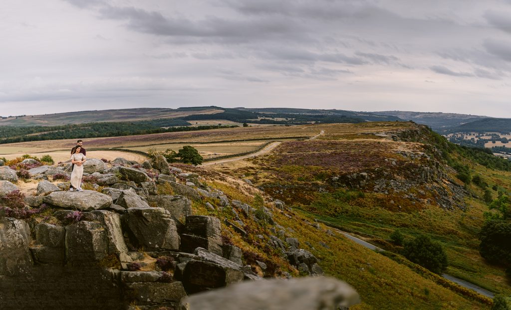 A person in a white outfit stands on rocks atop a grassy cliff at Curbar Edge, overlooking a vast landscape of rolling hills under a cloudy sky—a breathtaking setting for a memorable proposal.