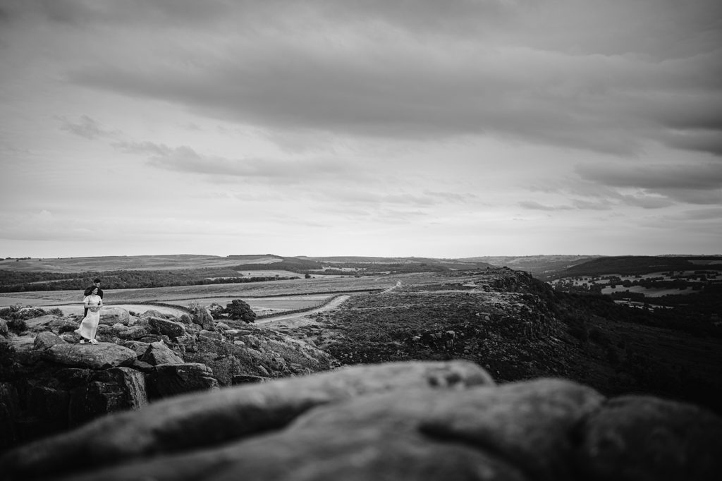 A person stands on a rocky outcrop at Curbar Edge, overlooking a vast landscape of hills and fields under a cloudy sky in black and white—a perfect setting for a proposal.