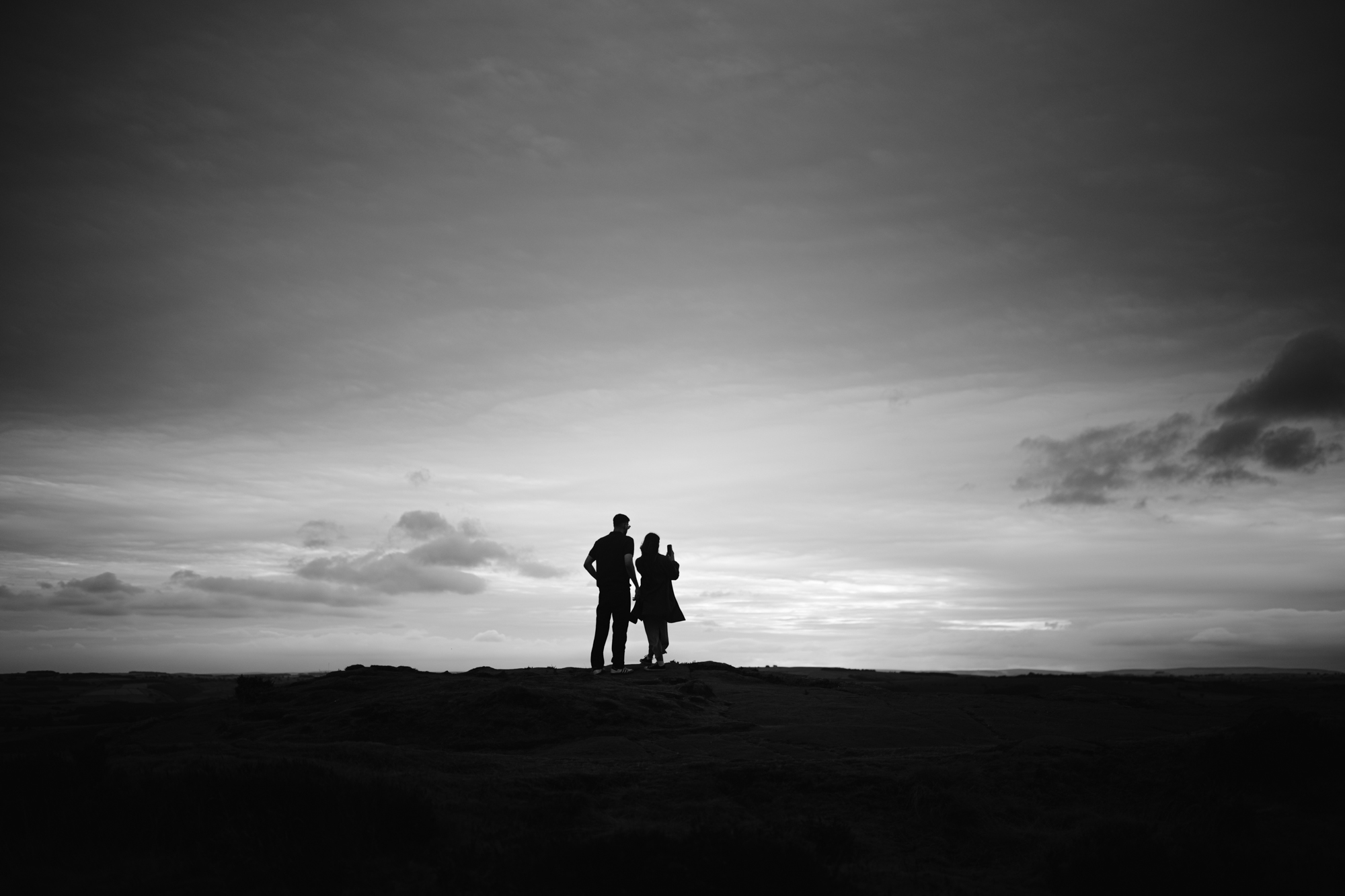 Silhouettes of two people standing together on a hilltop under a cloudy sky at sunset, capturing the magic of a Curbar Edge proposal.