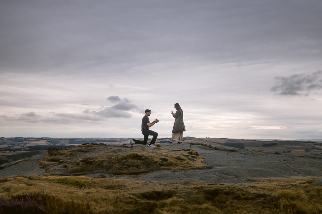A person kneels and proposes to another person on a rocky hilltop at Curbar Edge under a cloudy sky, with distant fields visible in the background.