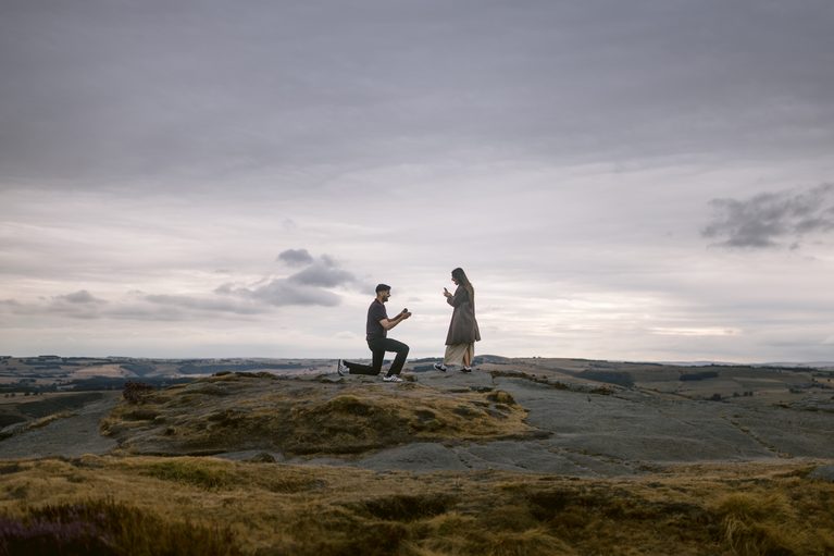 A person kneels and proposes to another person on a rocky hilltop at Curbar Edge under a cloudy sky, with distant fields visible in the background.