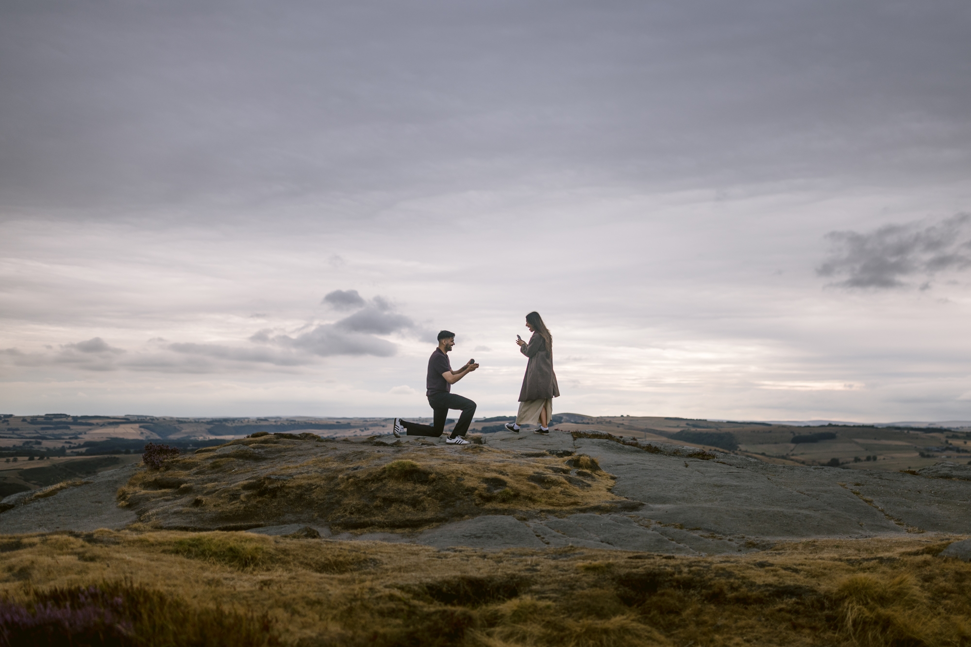 A person kneels and proposes to another person on a rocky hilltop at Curbar Edge under a cloudy sky, with distant fields visible in the background.