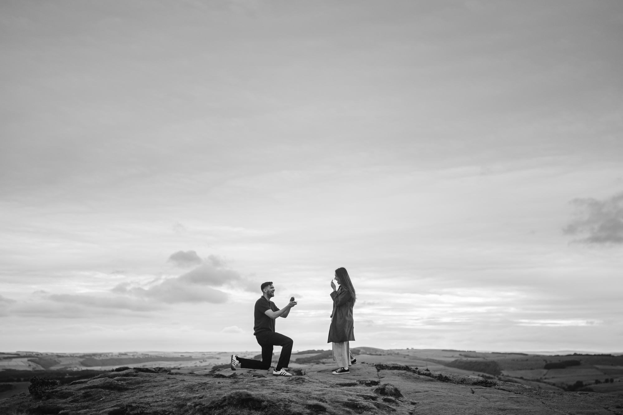 A person kneels and proposes to another person outdoors on rocky ground at Curbar Edge, with a wide sky and distant landscape in the background.