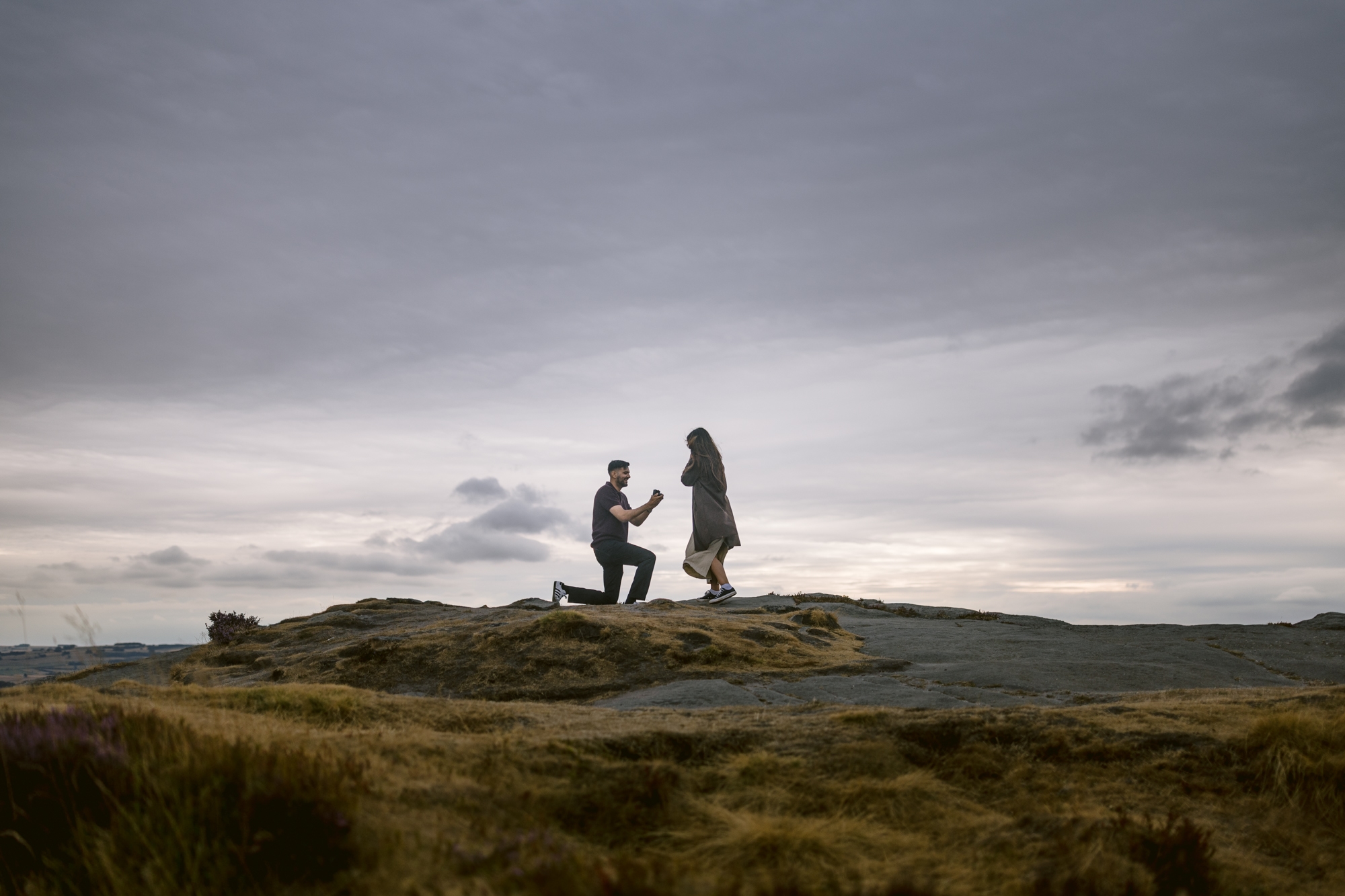 A person kneels and proposes to another on a rocky hilltop under a cloudy sky, with open landscape in the background, capturing the magic of a Curbar Edge proposal.