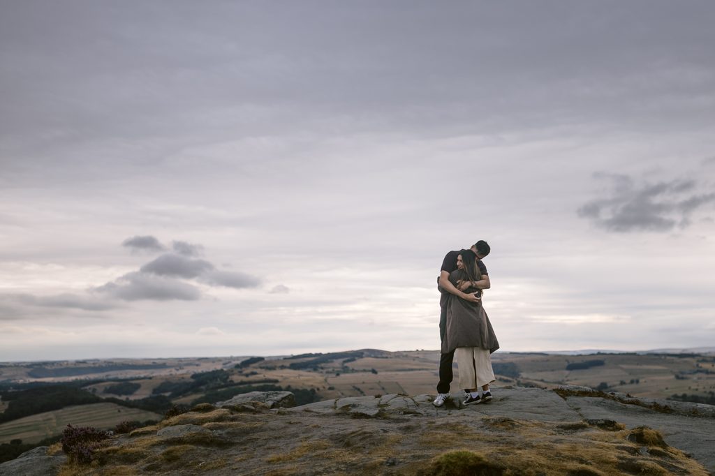 Two people embrace on a rocky hilltop under a cloudy sky, with rolling fields and distant hills visible in the background—capturing the magic of a Curbar Edge proposal.