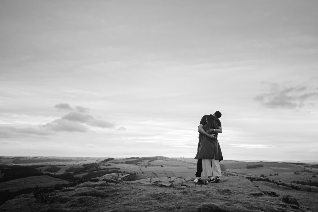 Two people embrace on a rocky hilltop at Curbar Edge, overlooking fields and distant hills under a cloudy sky in black and white—a moment reminiscent of a heartfelt proposal.
