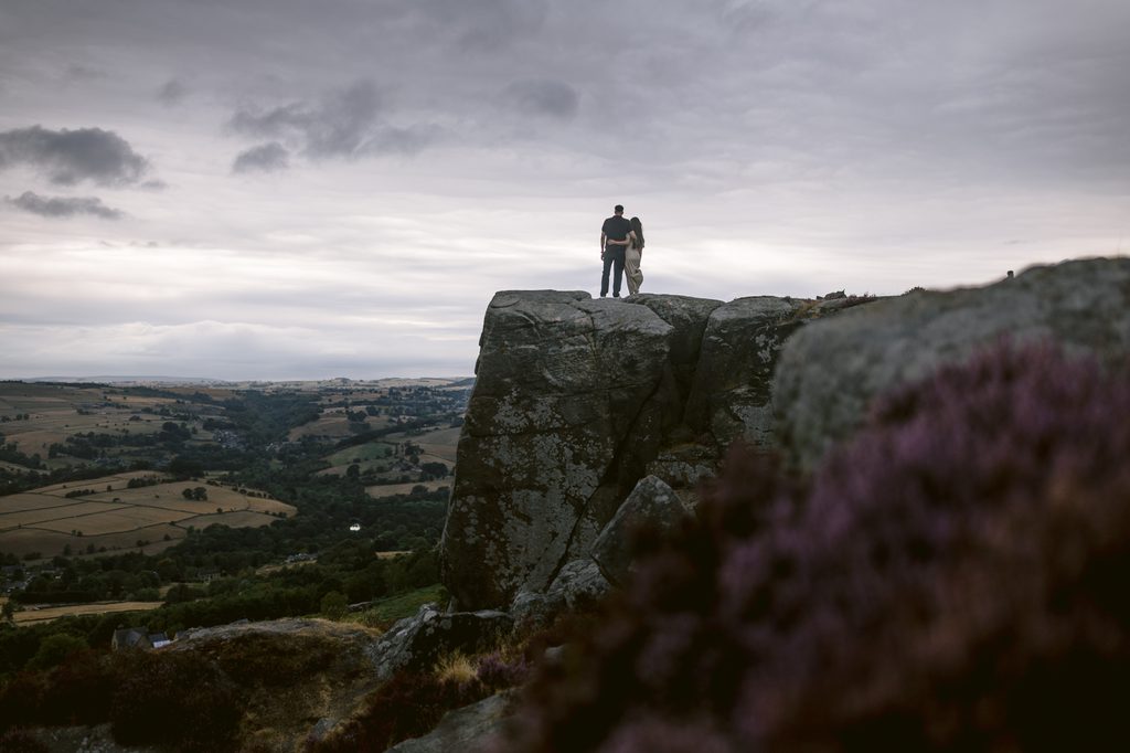Two people stand together on top of a large rock formation at Curbar Edge, overlooking a valley with fields under a cloudy sky—a breathtaking spot for a proposal.