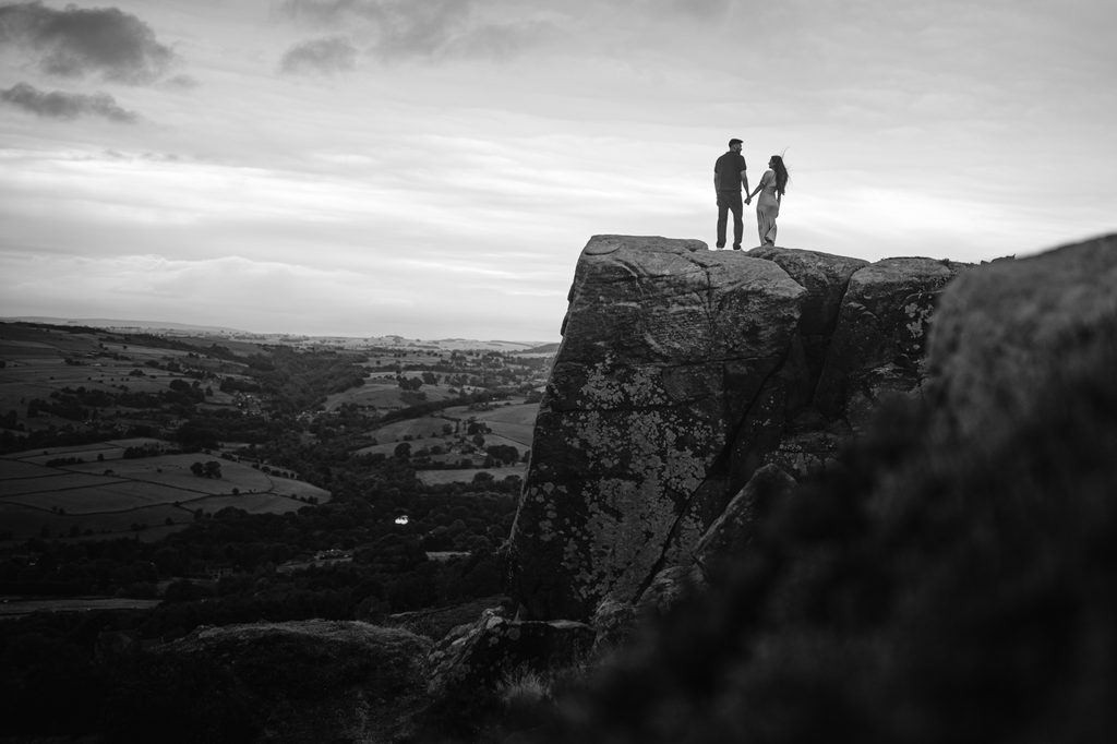 Two people stand holding hands on the edge of a large rocky cliff, possibly sharing a Curbar Edge proposal, as they overlook a landscape of hills and fields under a cloudy sky.