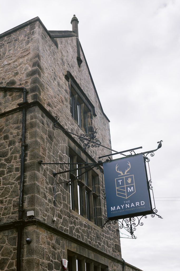 Stone building with rectangular windows, featuring a dark sign reading “The Maynard” with a crest and deer antlers, hanging from a black metal bracket—a perfect setting for a late summer wedding near Padley Gorge. Sky is overcast.