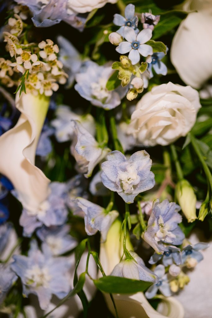 A close-up of a floral arrangement, perfect for a Late Summer Wedding at The Maynard, featuring pale blue delphiniums, white calla lilies, and small white blossoms with green foliage.