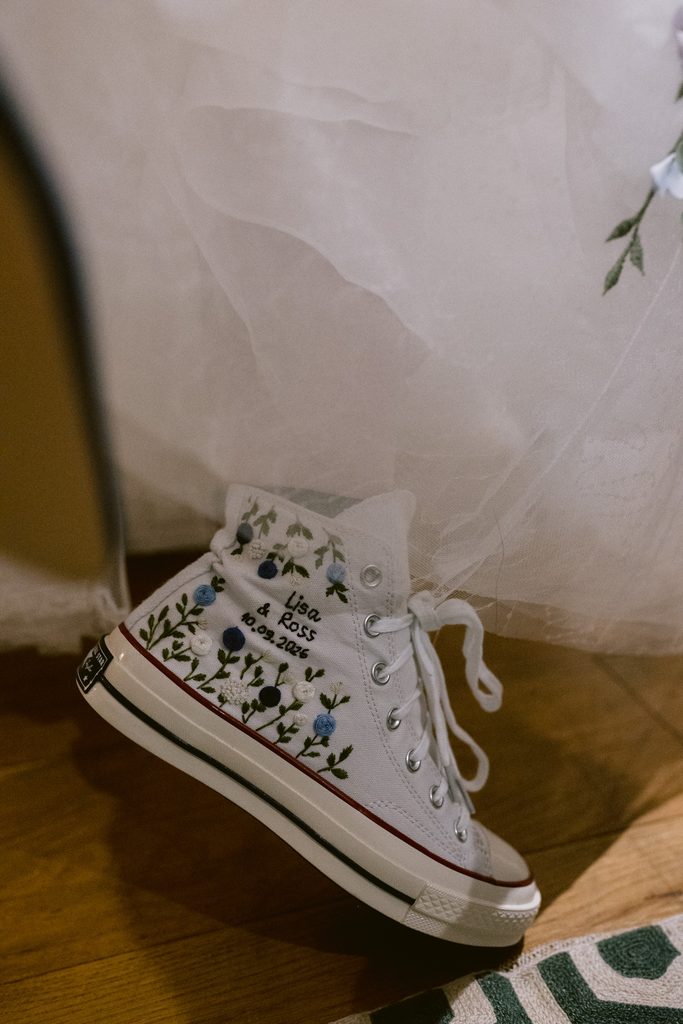 A white high-top trainer with floral designs and the text “Lisa & José 6.10.23” embroidered, partially covered by a sheer white fabric, rests on a wooden floor—a charming detail from a Late Summer Wedding at The Maynard near Padley Gorge.