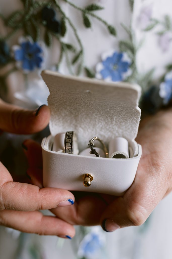 A person holding an open white ring box containing two rings, with blue floral embroidery visible in the background—the perfect detail for a Late Summer Wedding at The Maynard, nestled near the scenic beauty of Padley Gorge.