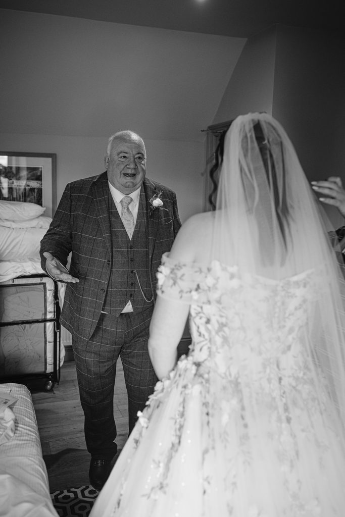 An older man in a suit and tie faces a bride in a wedding dress and veil inside a room at The Maynard, both appearing to share an emotional moment during their late summer wedding.
