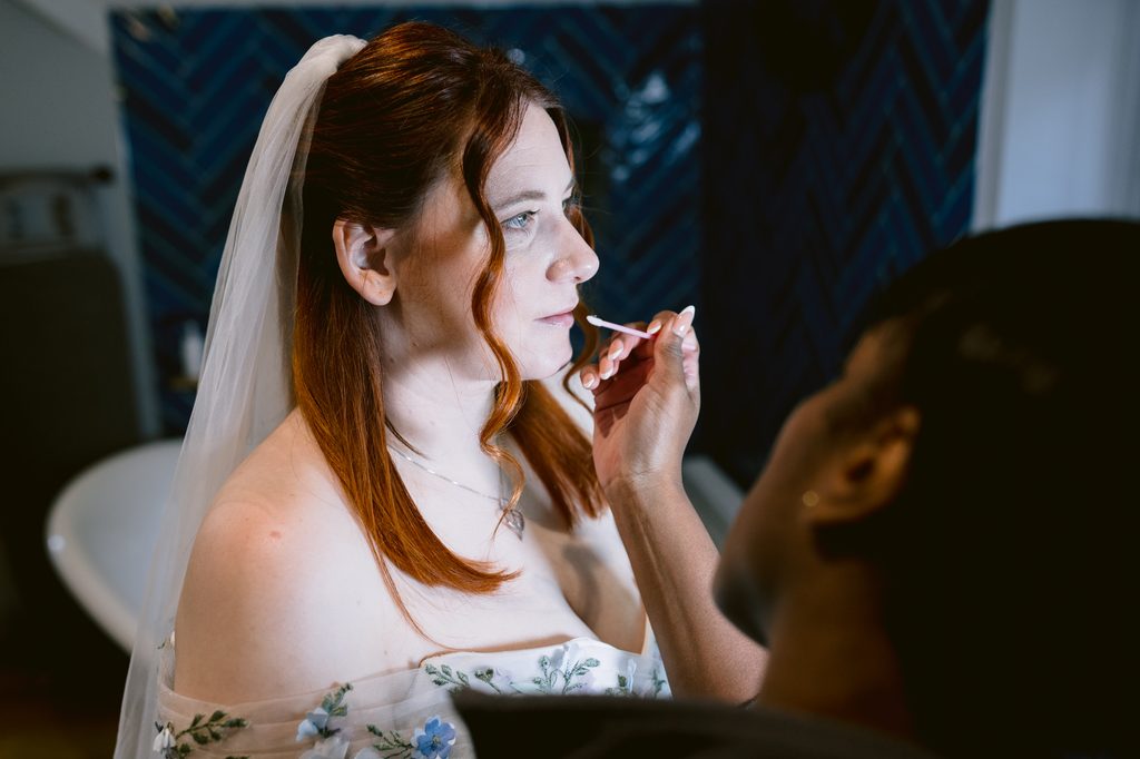 A bride with red hair and a veil has make-up applied by a make-up artist in a room with a blue tiled wall, preparing for her Late Summer Wedding at The Maynard.