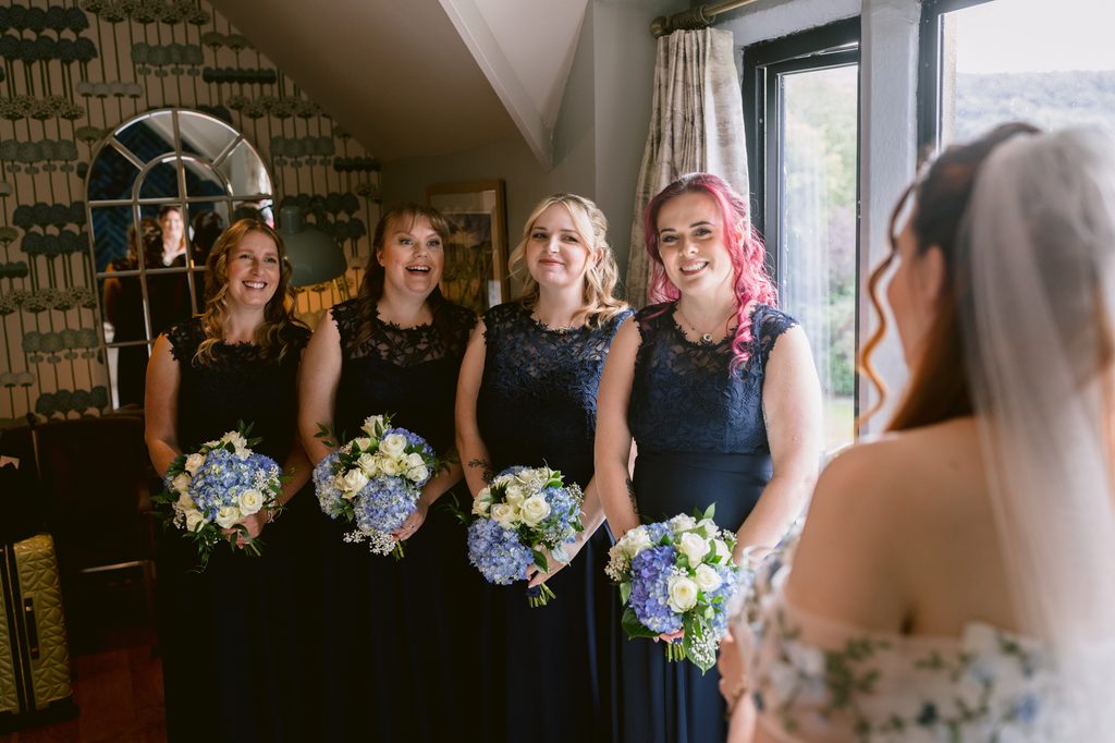 Four bridesmaids in matching navy dresses hold blue and white bouquets, smiling at a bride in a floral dress and veil inside a well-lit room with patterned wallpaper, capturing the joyful spirit of a Late Summer Wedding at The Maynard.