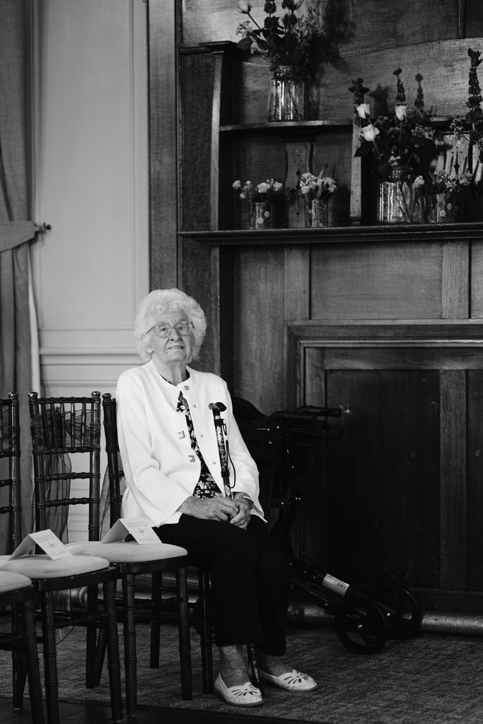 Elderly woman with white hair sits alone on a chair in a formal setting with wooden panelling and shelves with vases of flowers behind her, evoking the quiet elegance of a Late Summer Wedding at The Maynard.