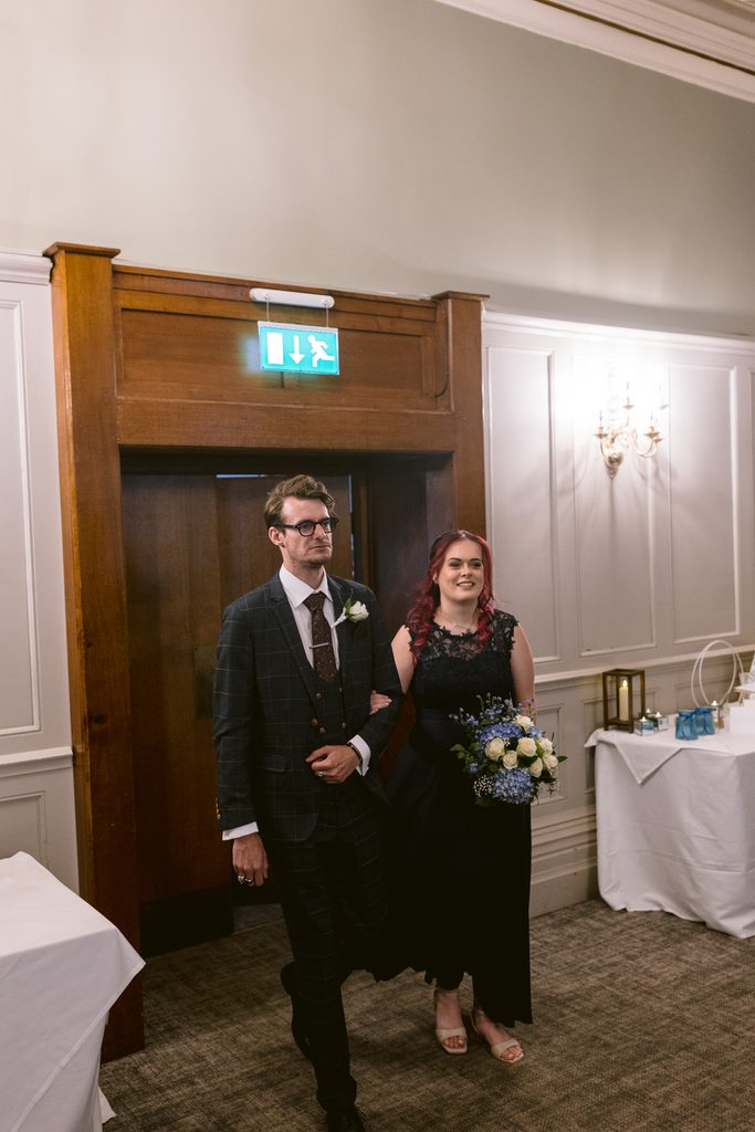 A man in a suit and a woman in a dark dress holding a bouquet walk arm in arm through a doorway at an indoor event, celebrating a late summer wedding at The Maynard.