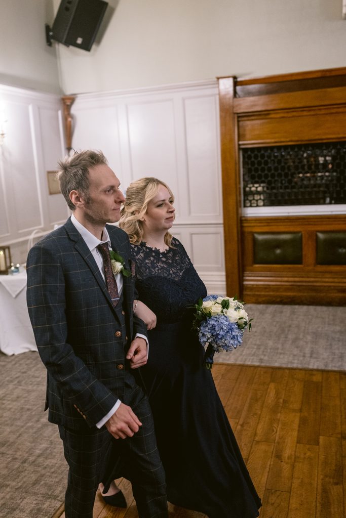 A man in a checked suit and a woman in a dark lace dress holding a bouquet walk side by side in a formal indoor setting, capturing the elegance of their Late Summer Wedding at The Maynard.