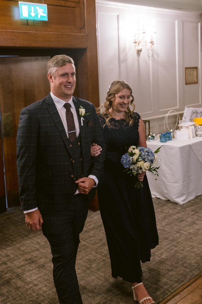 Man in a checked suit and woman in a navy dress holding a bouquet walk arm in arm indoors, smiling, during a formal event at The Maynard, capturing the joyful spirit of their late summer wedding.