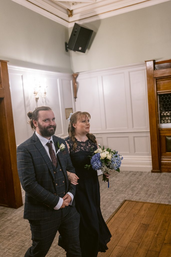 A man in a checked suit and a woman in a navy dress holding a bouquet walk arm-in-arm indoors at a formal event, capturing the elegant spirit of a late summer wedding at The Maynard.