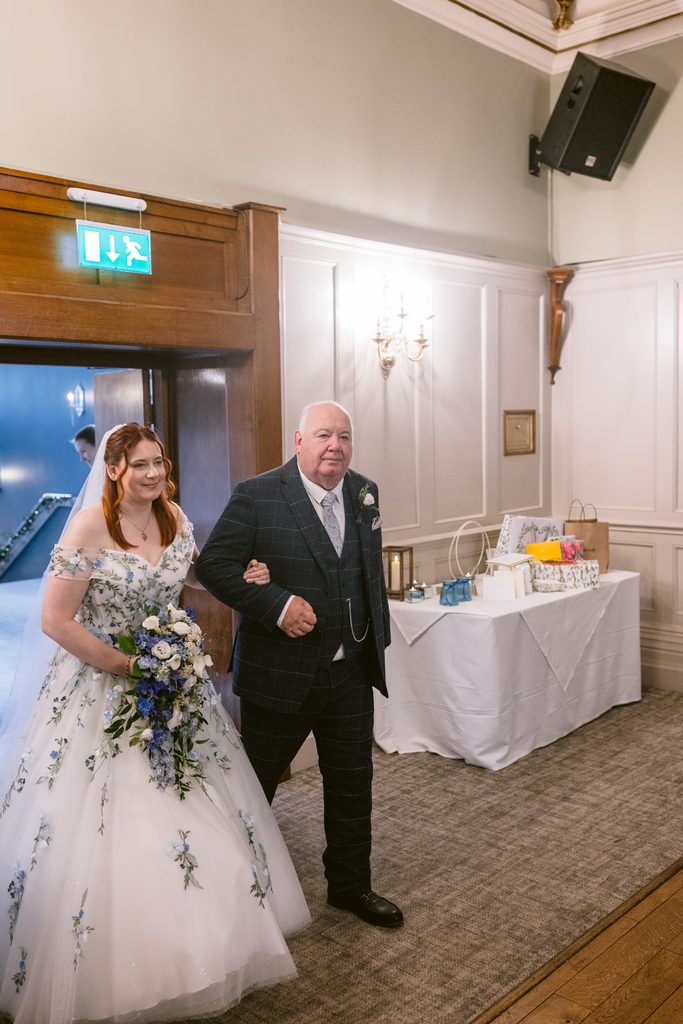 A bride in a white floral dress walks arm-in-arm with an older man in a suit, entering a room at The Maynard set up for a late summer wedding.