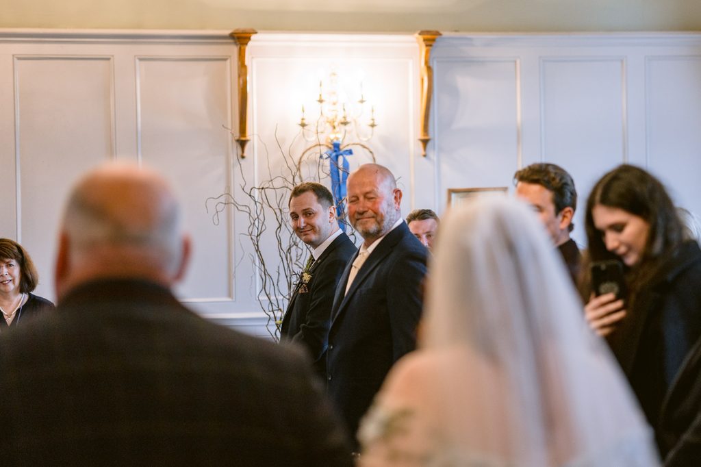 A groom smiles whilst standing beside an older man in a formal room at The Maynard, as a bride in a white dress approaches with guests watching during their late summer wedding.