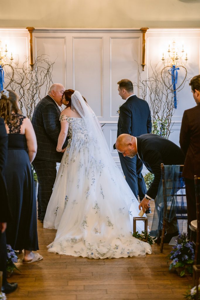 A bride and groom kiss at the altar during their late summer wedding ceremony at The Maynard, as guests stand nearby and a man adjusts a lantern on the floor.
