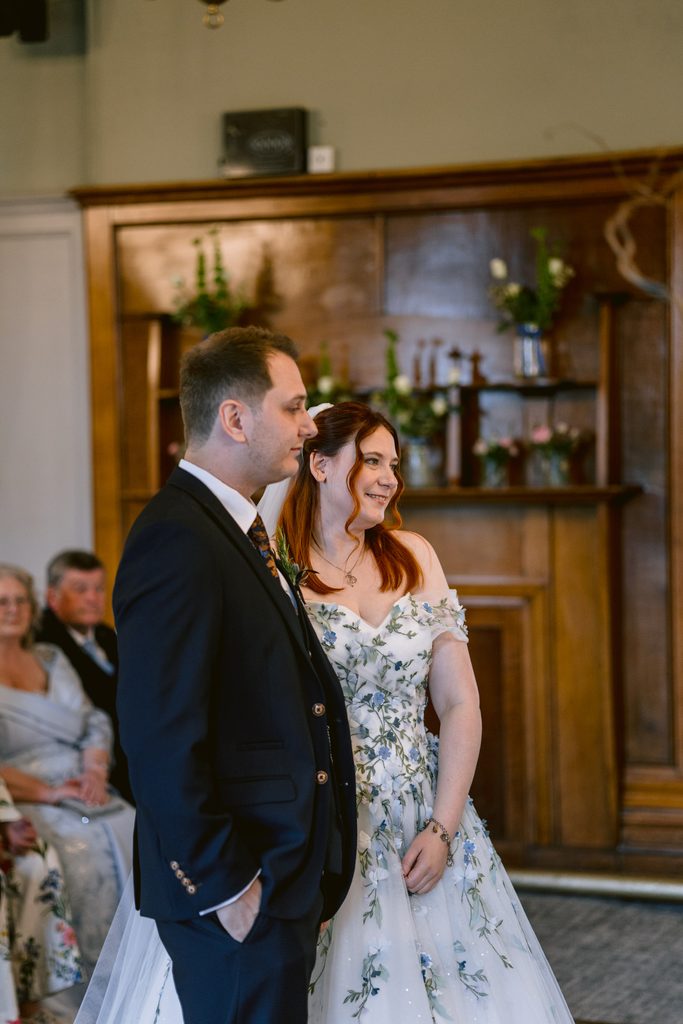A man in a suit and a woman in a white floral dress stand side by side indoors at The Maynard, with seated guests and a wooden cupboard in the background, capturing the elegant ambience of their late summer wedding.