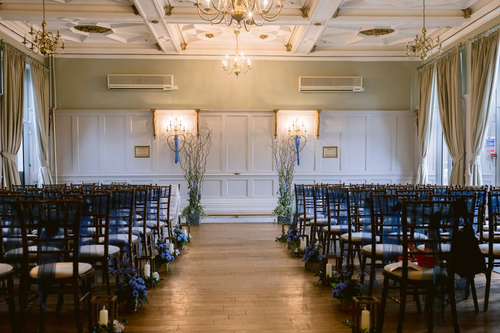 Rows of empty chairs face a decorated aisle in an elegant, light-filled room at The Maynard, set up for a late summer wedding ceremony, with floral arrangements along the aisle.
