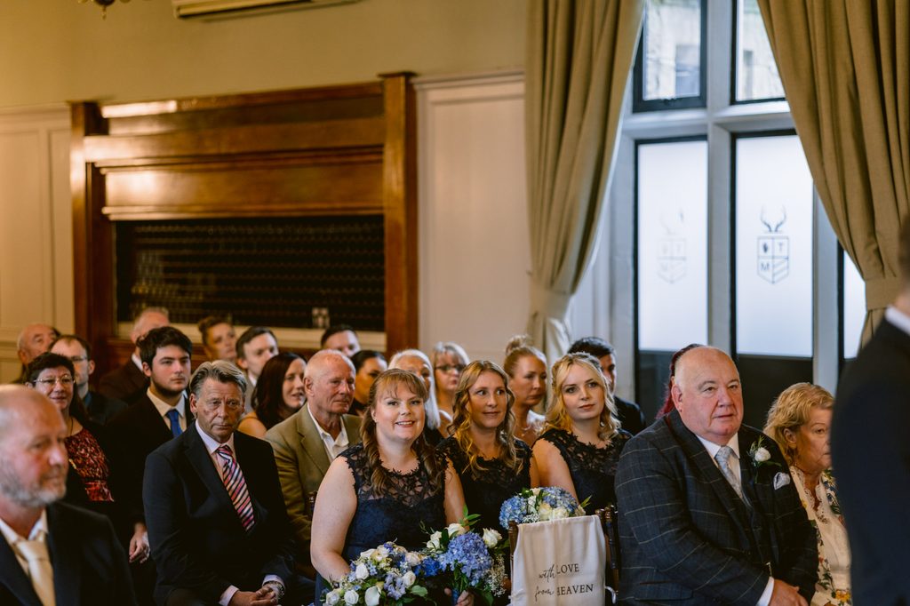 A group of people sit indoors at a Late Summer Wedding ceremony at The Maynard, with bridesmaids in navy dresses holding bouquets and guests looking forward attentively.