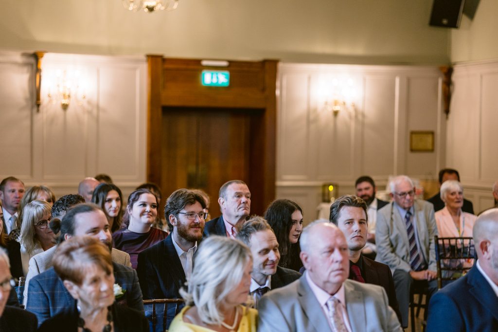 A group of people in formal attire sit in rows of chairs in a well-lit room with chandeliers and wood panelling, facing forward, as they eagerly await the start of a late summer wedding at The Maynard.