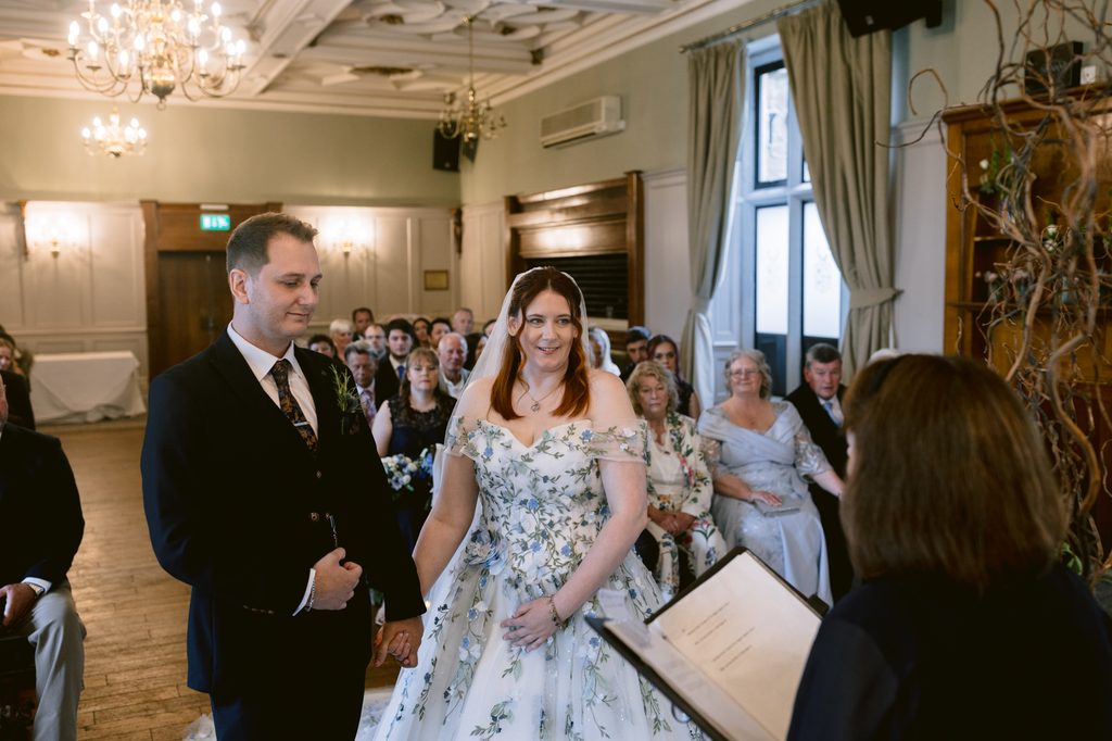 A couple stands at the altar holding hands during a Late Summer Wedding ceremony in a beautifully decorated room at The Maynard, with guests seated and an officiant reading from a book.